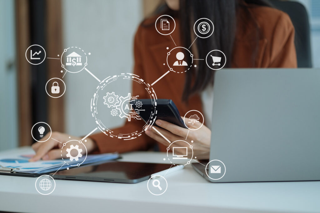 Op de afbeelding is een watermerk te zien van AI, met icoontjes waar AI op van toepassing is. En op de achtergrond is een vrouw te zien die achter haar laptop zit en wat documenten doorleest, en een telefoon in haar hand heeft.